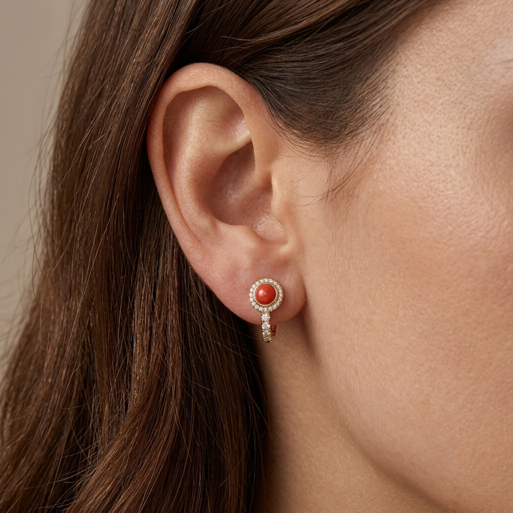 A close-up of a model's ear wearing a delicate gold dangle earring with a round coral cabochon and small clear accent stones.