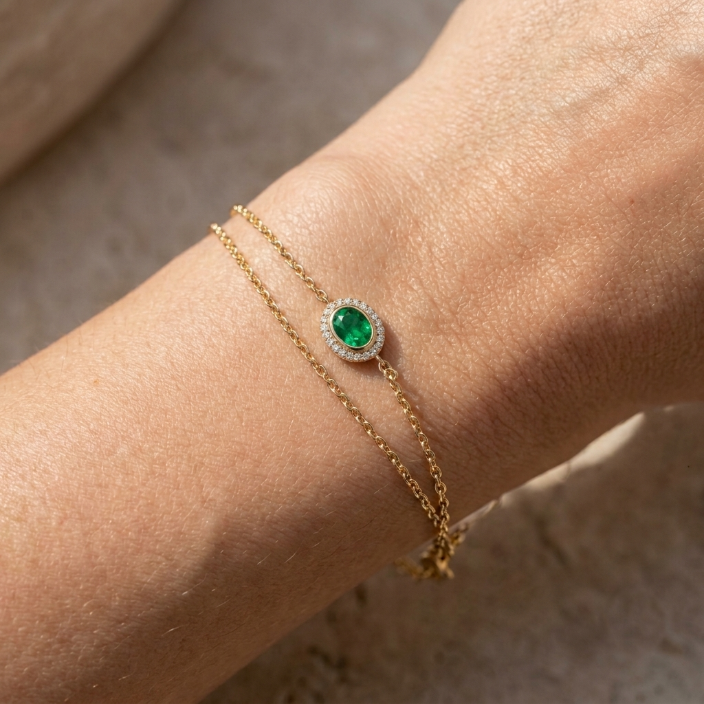 A close-up of a gold double-strand bracelet worn on a wrist, featuring a central oval green emerald stone surrounded by small clear stones.