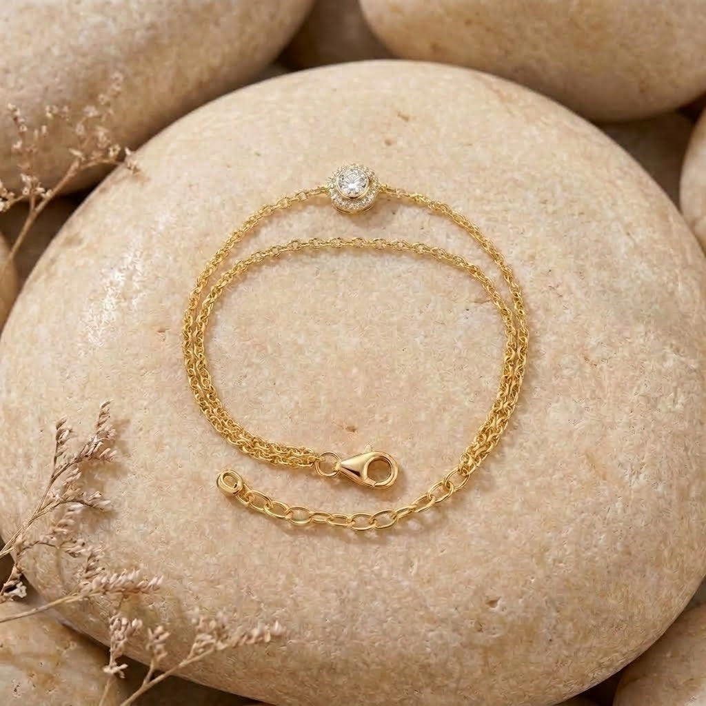A delicate gold chain bracelet with a single round clear gemstone set in a bezel, displayed on a smooth beige stone with dried floral accents.