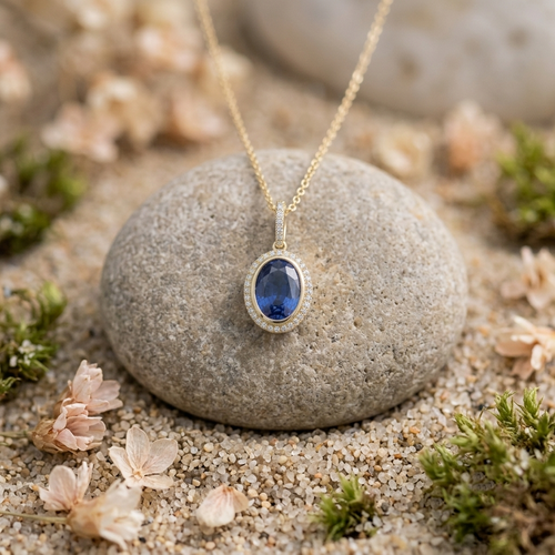 A gold necklace with an oval blue gemstone pendant, displayed on a smooth gray stone surrounded by sand, small flowers, and moss.