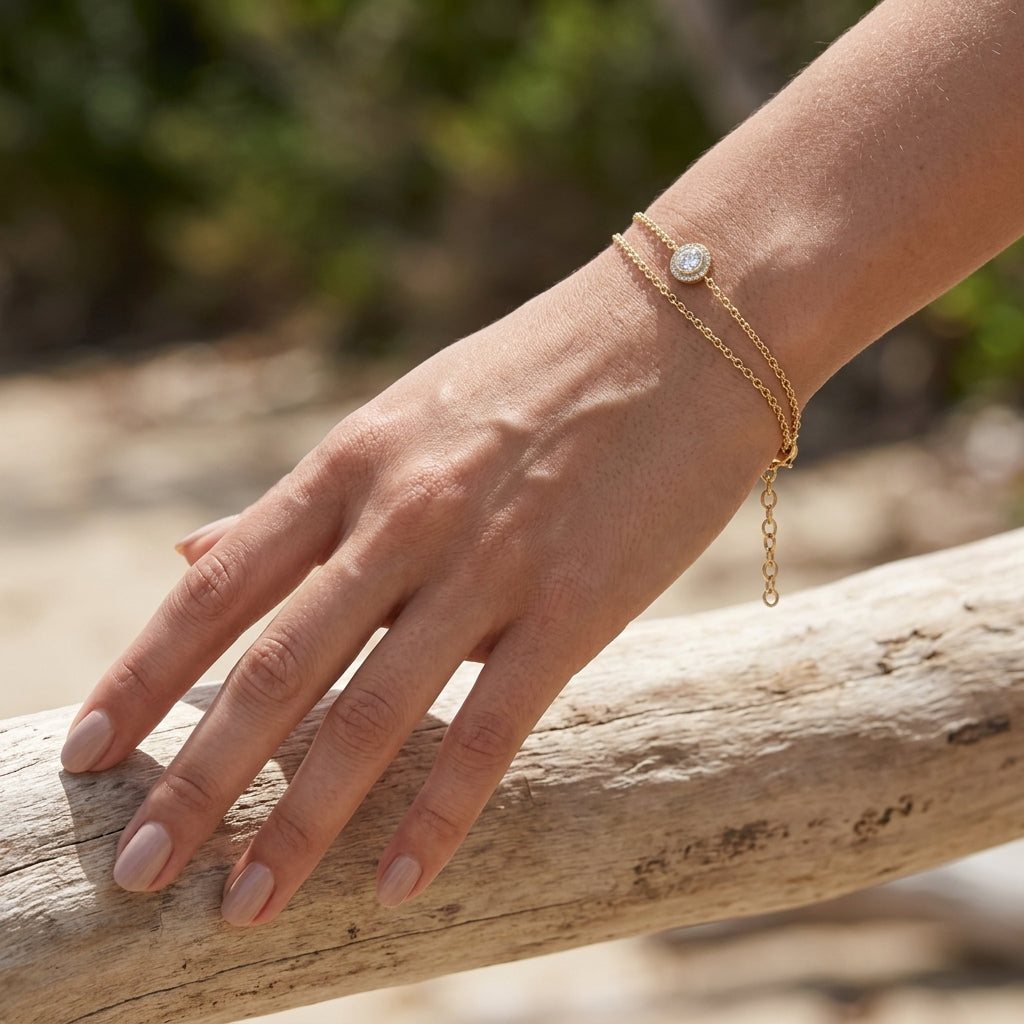 A close-up of a person's hand resting on a weathered log outdoors, wearing a delicate gold double-strand bracelet with a small round clear stone.