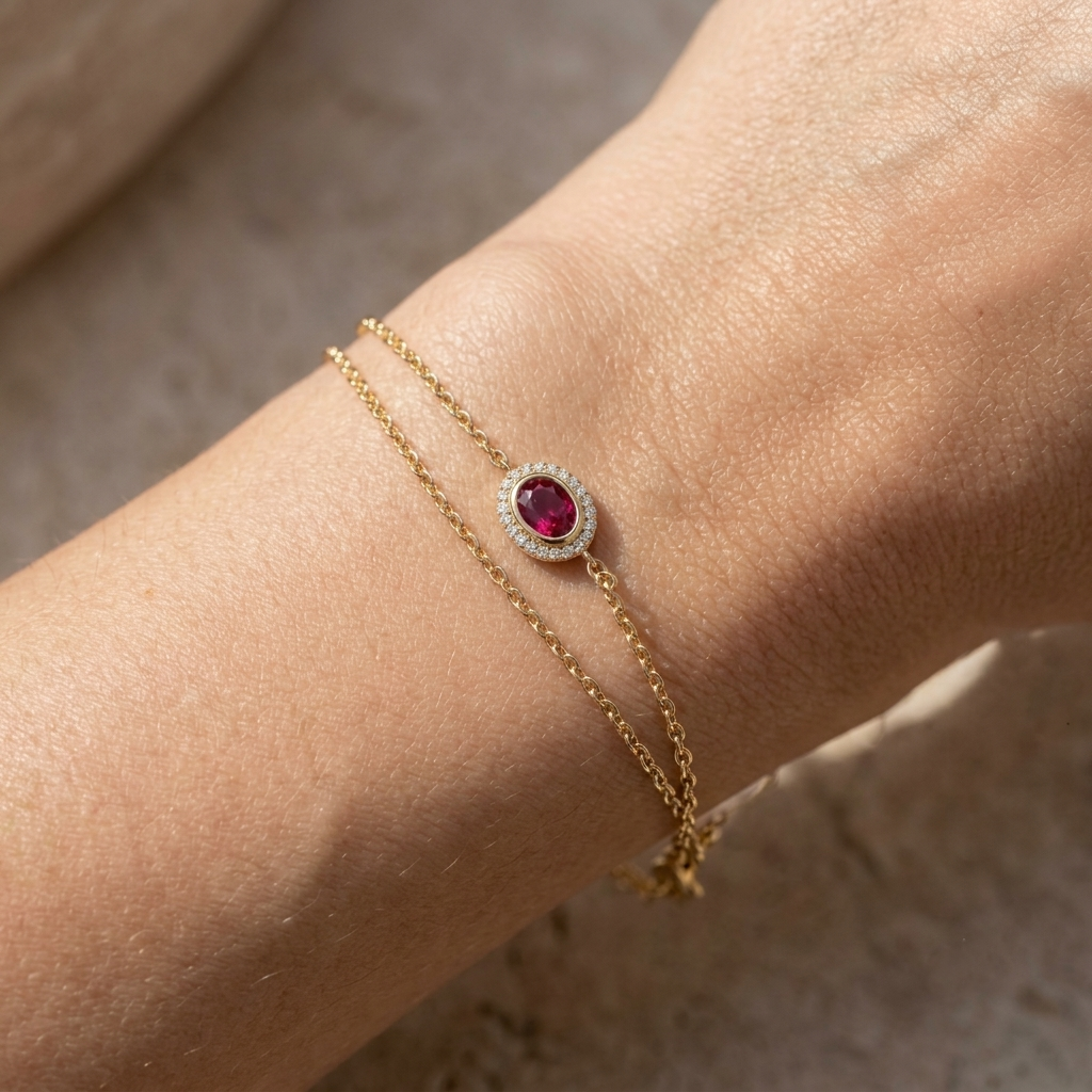 A close-up of a delicate gold double-strand bracelet worn on a wrist, featuring a central oval ruby gemstone surrounded by a halo of small clear stones.