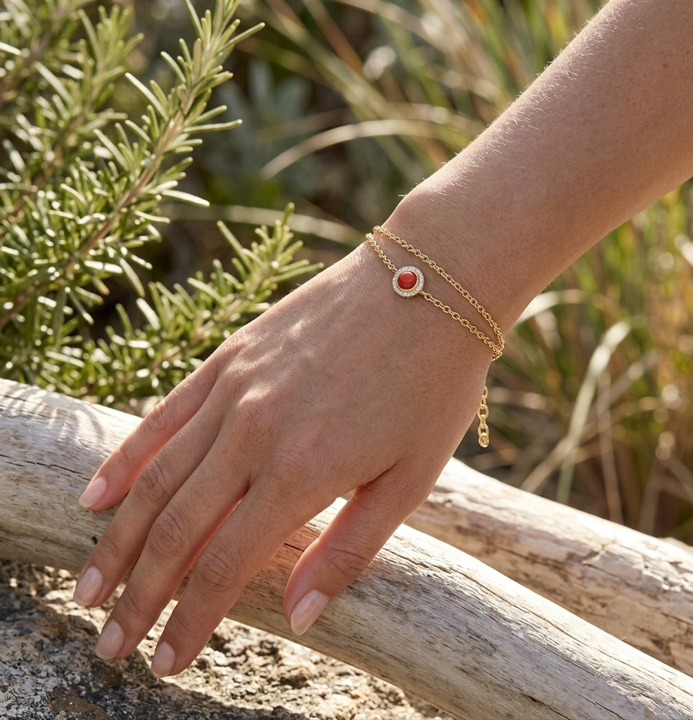 A close-up of a person's hand resting on a log outdoors, wearing a delicate double-strand gold chain bracelet with a small red and white circular charm.